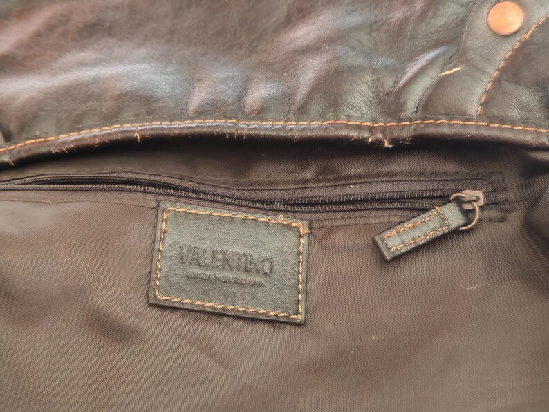 Peut inclure: L'int&eacute;rieur d'un sac en cuir marron fonc&eacute; avec une &eacute;tiquette rectangulaire en cuir portant l'inscription VALENTINO en relief. La doublure est en tissu marron fonc&eacute; avec une poche zipp&eacute;e horizontale, une tirette en cuir et des coutures orange.