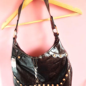 Peut inclure: Un sac &agrave; bandouli&egrave;re marron fonc&eacute; &agrave; la finition brillante, dot&eacute; d'une bordure festonn&eacute;e orn&eacute;e de clous m&eacute;talliques sur le devant. Le sac comprend une bandouli&egrave;re unique, un d&eacute;tail de cha&icirc;ne argent&eacute;e au centre et une petite &eacute;tiquette produit.