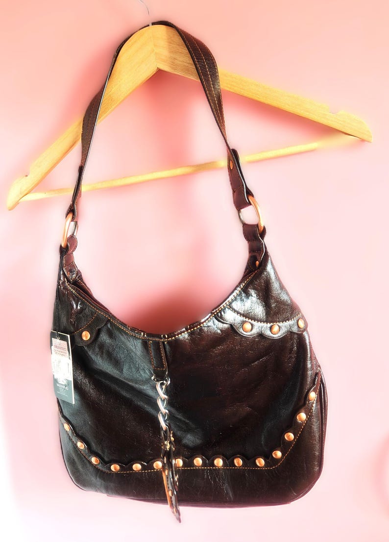 Peut inclure: Un sac &agrave; bandouli&egrave;re marron fonc&eacute; &agrave; la finition brillante, dot&eacute; d'une bordure festonn&eacute;e orn&eacute;e de clous m&eacute;talliques sur le devant. Le sac comprend une bandouli&egrave;re unique, un d&eacute;tail de cha&icirc;ne argent&eacute;e au centre et une petite &eacute;tiquette produit.