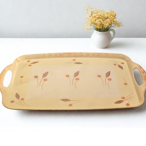 Könnte beinhalten: Ein rechteckiges Serviertablett mit Griffen. Das Tablett hat einen hellgelben Hintergrund mit einem floralen Muster in Braun- und Orangetönen. Die Ränder und Griffe haben eine dunklere, strukturierte Oberfläche. Im Hintergrund steht eine kleine weiße Vase mit gelben Blumen.