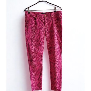 May include: A pair of pink snake print pants hanging on a black hanger against a white background. The pants have a button and zipper closure, and pockets. The snake print pattern is a mix of dark and light pink.
