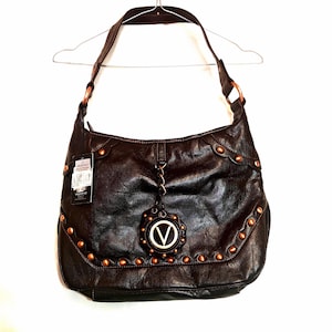 Peut inclure: Un sac hobo marron fonc&eacute; avec un bord sup&eacute;rieur festonn&eacute;, des clous m&eacute;talliques couleur cuivre sur le devant et une breloque circulaire centrale avec un logo V blanc. Le sac comprend une bandouli&egrave;re fix&eacute;e par des anneaux en cuivre et une &eacute;tiquette.