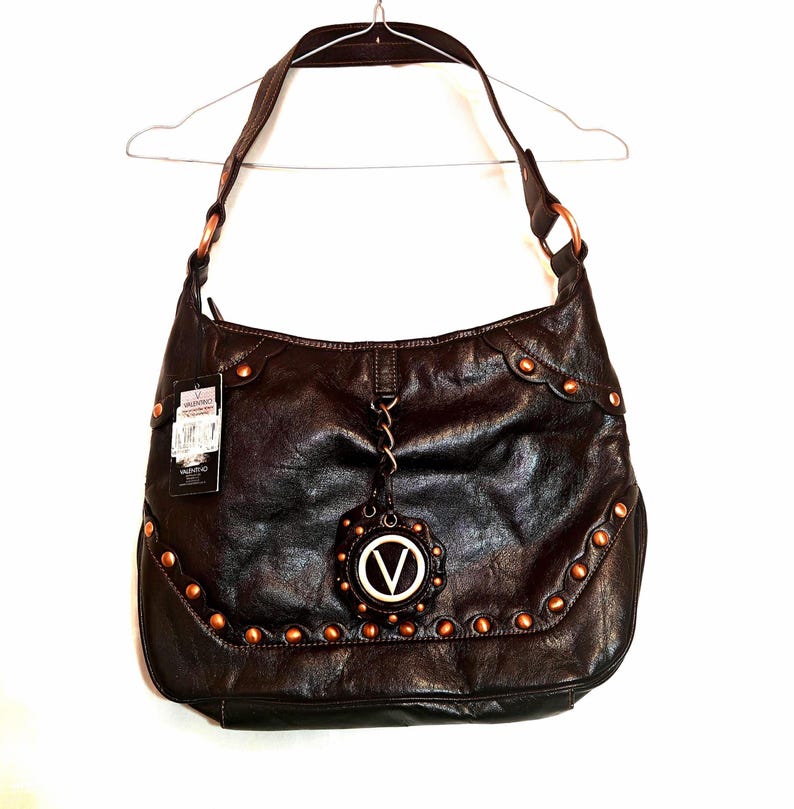 Peut inclure: Un sac hobo marron fonc&eacute; avec un bord sup&eacute;rieur festonn&eacute;, des clous m&eacute;talliques couleur cuivre sur le devant et une breloque circulaire centrale avec un logo V blanc. Le sac comprend une bandouli&egrave;re fix&eacute;e par des anneaux en cuivre et une &eacute;tiquette.
