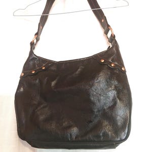 Peut inclure: Un sac hobo en cuir marron fonc&eacute; dot&eacute; d'une bandouli&egrave;re unique fix&eacute;e par des anneaux m&eacute;talliques circulaires couleur cuivre. Le bord sup&eacute;rieur du sac pr&eacute;sente une bordure festonn&eacute;e accentu&eacute;e par de petits clous m&eacute;talliques cuivr&eacute;s.