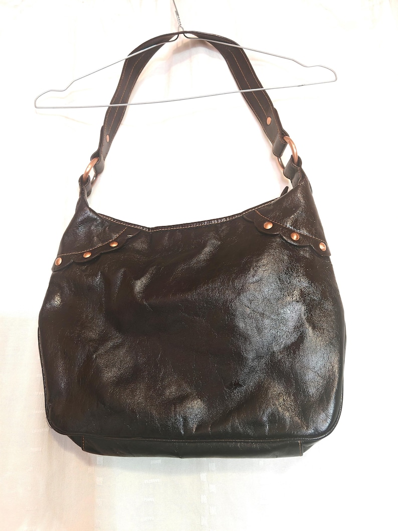 Peut inclure: Un sac hobo en cuir marron fonc&eacute; dot&eacute; d'une bandouli&egrave;re unique fix&eacute;e par des anneaux m&eacute;talliques circulaires couleur cuivre. Le bord sup&eacute;rieur du sac pr&eacute;sente une bordure festonn&eacute;e accentu&eacute;e par de petits clous m&eacute;talliques cuivr&eacute;s.