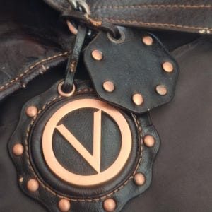 Peut inclure: Un sac marron fonc&eacute; dot&eacute; d'une breloque en cuir d&eacute;corative avec un embl&egrave;me circulaire cuivr&eacute; affichant la lettre V. La breloque est orn&eacute;e de petits clous en cuivre et est fix&eacute;e au sac par une cha&icirc;ne en m&eacute;tal fonc&eacute; et une lani&egrave;re en cuir.