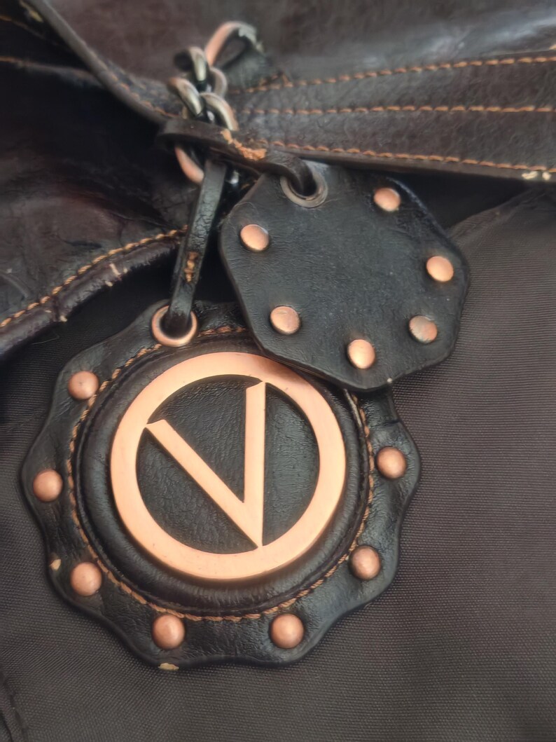 Peut inclure: Un sac marron fonc&eacute; dot&eacute; d'une breloque en cuir d&eacute;corative avec un embl&egrave;me circulaire cuivr&eacute; affichant la lettre V. La breloque est orn&eacute;e de petits clous en cuivre et est fix&eacute;e au sac par une cha&icirc;ne en m&eacute;tal fonc&eacute; et une lani&egrave;re en cuir.