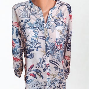 Può includere: Una camicia abbottonata di colore chiaro con un motivo floreale. La camicia presenta uno scollo a V e maniche lunghe. Il design floreale comprende foglie e fiori blu, rossi e verdi su uno sfondo chiaro.