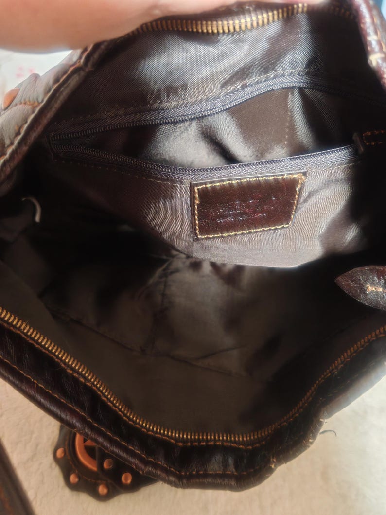 Peut inclure: L'int&eacute;rieur d'un sac &agrave; main marron fonc&eacute; avec une doublure en tissu marron fonc&eacute;, une fermeture &eacute;clair sur le dessus et une poche int&eacute;rieure zipp&eacute;e. Un &eacute;cusson en cuir marron fonc&eacute; est cousu sur la doublure avec du texte en relief.