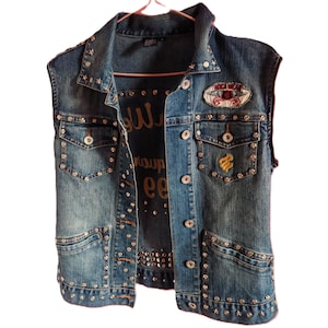May include: A blue denim vest featuring silver-toned metal studs along the collar, pockets, and hem. The garment includes two chest pockets with button closures, a yellow embroidered logo on the right pocket, and a patch on the upper left chest.