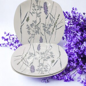 Op de afbeelding: Twee witte keramische borden met een vierkante vorm en afgeronde hoeken. De borden hebben een delicaat bloemmotief in tinten paars, groen en grijs. Het ontwerp omvat hoog gras en lavendel takjes. Een boeket paarse bloemen staat op de achtergrond.