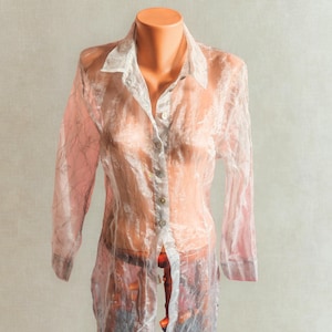 May include: A translucent, pale pink button-up shirt with long sleeves, shown on a mannequin. The shirt features a white collar and cuffs, and a delicate, textured design. The buttons are small and pearlescent.