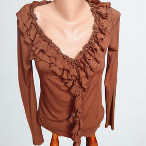 May include: A brown long-sleeve top with a deep V-neckline and ruffled trim. The ruffles cascade down the front and around the neckline. The top is displayed on a mannequin.