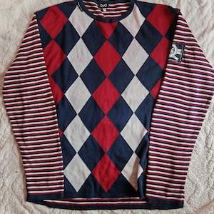 Può includere: Un maglione a maniche lunghe con un motivo a rombi rosso, bianco e blu. Le maniche sono a righe rosse, bianche e blu. Il maglione ha un logo D&G sul davanti.