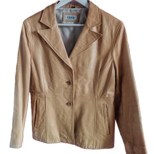 May include: A tan leather jacket with a classic design. The jacket features a notched collar, two front pockets, and a two-button closure. The jacket is hanging on a white hanger.