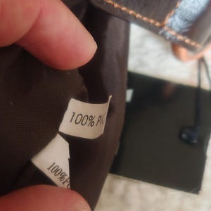 Peut inclure: L'int&eacute;rieur d'un sac marron fonc&eacute; avec une &eacute;tiquette d'entretien en tissu blanc portant l'inscription 100% PVC en noir. Le sac pr&eacute;sente une doublure marron fonc&eacute; et un rabat ext&eacute;rieur marron fonc&eacute; avec des coutures orange visibles sur le bord.