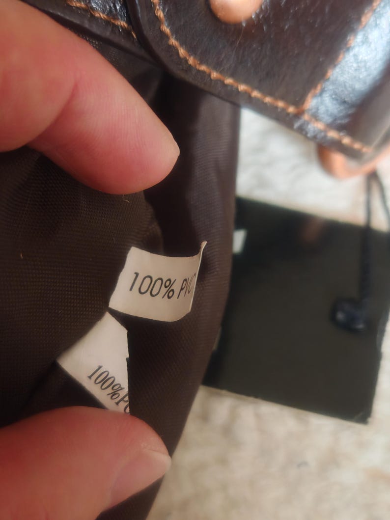 Peut inclure: L'int&eacute;rieur d'un sac marron fonc&eacute; avec une &eacute;tiquette d'entretien en tissu blanc portant l'inscription 100% PVC en noir. Le sac pr&eacute;sente une doublure marron fonc&eacute; et un rabat ext&eacute;rieur marron fonc&eacute; avec des coutures orange visibles sur le bord.