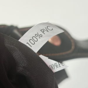 Peut inclure: Un v&ecirc;tement noir dot&eacute; d'une &eacute;tiquette d'entretien en tissu blanc avec le texte 100% PVC imprim&eacute; en noir. Le mat&eacute;riau pr&eacute;sente une texture en maille fine, et une deuxi&egrave;me &eacute;tiquette blanche partiellement visible affiche &eacute;galement 100% PVC.