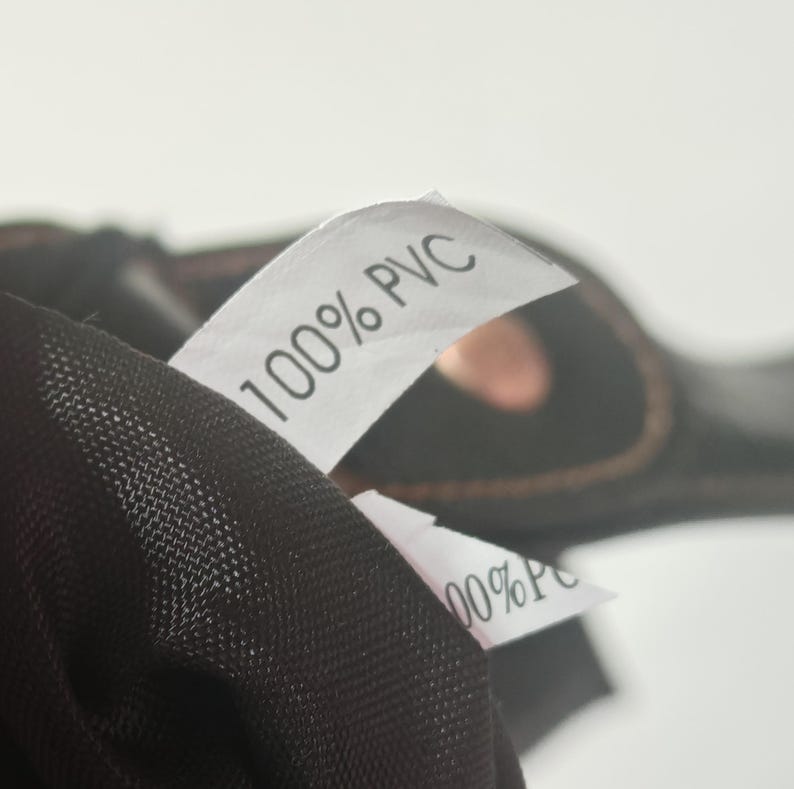 Peut inclure: Un v&ecirc;tement noir dot&eacute; d'une &eacute;tiquette d'entretien en tissu blanc avec le texte 100% PVC imprim&eacute; en noir. Le mat&eacute;riau pr&eacute;sente une texture en maille fine, et une deuxi&egrave;me &eacute;tiquette blanche partiellement visible affiche &eacute;galement 100% PVC.