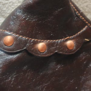 Peut inclure: Un accessoire en cuir marron fonc&eacute; pr&eacute;sentant une bordure festonn&eacute;e le long de la couture sup&eacute;rieure. La section festonn&eacute;e est orn&eacute;e de trois clous m&eacute;talliques circulaires de couleur cuivre et fix&eacute;e par des coutures orange-brun visibles.