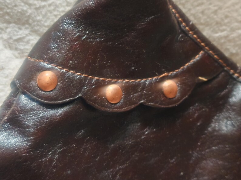 Peut inclure: Un accessoire en cuir marron fonc&eacute; pr&eacute;sentant une bordure festonn&eacute;e le long de la couture sup&eacute;rieure. La section festonn&eacute;e est orn&eacute;e de trois clous m&eacute;talliques circulaires de couleur cuivre et fix&eacute;e par des coutures orange-brun visibles.