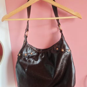 Peut inclure: Un sac &agrave; bandouli&egrave;re de style hobo marron fonc&eacute; avec une finition brillante. Le sac pr&eacute;sente une large sangle unique fix&eacute;e par des anneaux m&eacute;talliques circulaires couleur cuivre. Une bordure festonn&eacute;e d&eacute;core le haut, fix&eacute;e par des rivets cuivr&eacute;s.