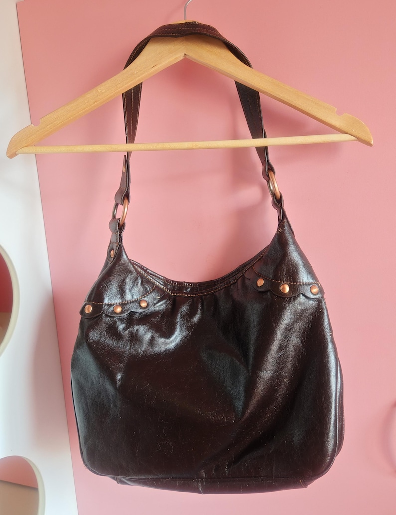 Peut inclure: Un sac &agrave; bandouli&egrave;re de style hobo marron fonc&eacute; avec une finition brillante. Le sac pr&eacute;sente une large sangle unique fix&eacute;e par des anneaux m&eacute;talliques circulaires couleur cuivre. Une bordure festonn&eacute;e d&eacute;core le haut, fix&eacute;e par des rivets cuivr&eacute;s.