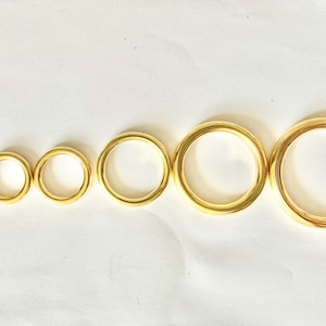 May include: Six gold-coloured metal rings of varying sizes are arranged in a row. The rings are smooth and shiny, with a consistent golden hue. The sizes range from small to large, showcasing a graduated scale.