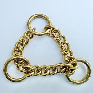May include: A gold-coloured metal chain accessory featuring three circular rings and a triangular chain design. The chain links are thick and interlocked, creating a bold, geometric pattern. Likely a fashion accessory.