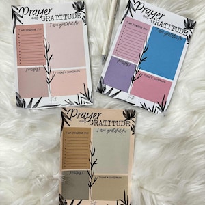 Op de afbeelding: Drie gebeds- en dankbaarheidsdagboeken met bloemmotieven. Elk dagboek heeft secties voor gebeden, dankbaarheid en schrift. De dagboeken hebben pastelkleurige blokken en de tekst "Prayer and Gratitude". Een pen ligt tussen twee dagboeken.