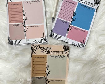 May include: Three rectangular paper notepads titled Prayer and GRATITUDE with sections for I am praying for, I am grateful for, praises, and Today's Scripture. Each notepad features black botanical illustrations and a white pen is included.