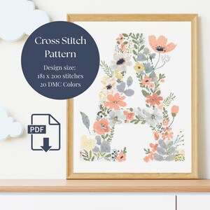 May include: Cross stitch pattern of the letter "A" decorated with peach, white, and blue flowers. The design size is 181 x 200 stitches, using 20 DMC colours. The framed artwork is displayed on a wooden shelf.