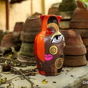 May include: A terracotta vase with a hand-painted design of a woman's face with orange, red, gold, and black accents.