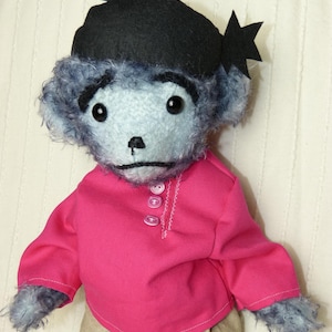 May include: A blue teddy bear wearing a black hat with a star on top, a pink shirt with three buttons, and beige pants.