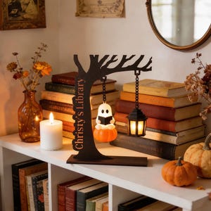 May include: A dark brown wooden tree-shaped display stand with the text 'Christy's Library' laser-cut into the trunk. It features two hanging elements: a small white ghost figure sitting on an orange pumpkin and a black lantern with a warm light.