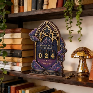 May include: A gothic-arched wooden book tracker featuring a purple and blue night sky illustration with mountains, clouds, and stars. It displays the text "BOOKS READ THIS YEAR" and "CHRISTY" with three rotating number tiles currently set to 024.