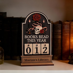 Könnte beinhalten: Ein hölzerner Bücherzähler mit schwarzem Hintergrund, mit einem Totenkopf, Rosen und dem Text "BOOKS READ THIS YEAR". Der Zähler zeigt "012" an und ist mit "Marian's Library" personalisiert.