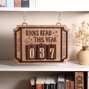 Könnte beinhalten: Holzschild mit der Aufschrift "BOOKS READ THIS YEAR" und der Zahl "037". Das Schild, in Form eines Tickets, dient zur Verfolgung der gelesenen Bücher. Es hängt an einem Regal.