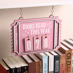 Könnte beinhalten: Ein rosa Holzschild mit der Aufschrift "BOOKS READ THIS YEAR" und der Zahl "078". Das Schild hängt über einer Sammlung von Büchern. Das Schild zeigt auch "ADMIT ONE" und "READER ID: HANNA".