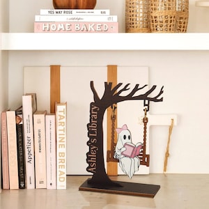 May include: A wooden decorative piece featuring a tree silhouette with a swing. A cartoon ghost with a pink bow reads a book on the swing. The text "Ashley's Library" is on the tree trunk. Books and decorative items are on the shelves.