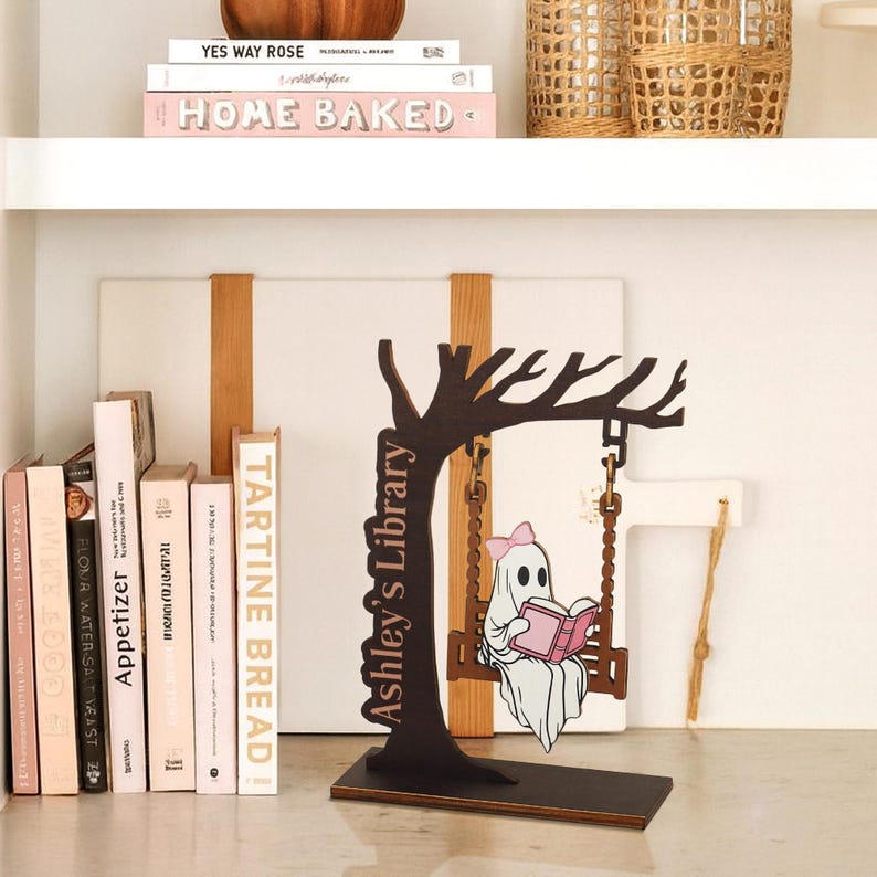 May include: A wooden decorative piece featuring a tree silhouette with a swing. A cartoon ghost with a pink bow reads a book on the swing. The text "Ashley's Library" is on the tree trunk. Books and decorative items are on the shelves.