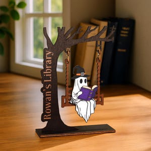 May include: A wooden Halloween decoration featuring a ghost in a witch hat reading a purple book while swinging on a swing. The swing is suspended from a tree-shaped structure. The text "Rowan's Library" is on the tree trunk.