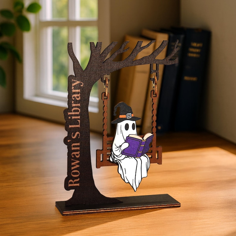 May include: A wooden Halloween decoration featuring a ghost in a witch hat reading a purple book while swinging on a swing. The swing is suspended from a tree-shaped structure. The text "Rowan's Library" is on the tree trunk.