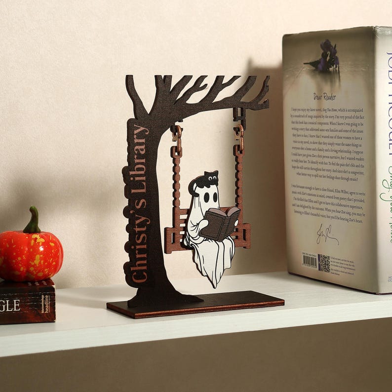 May include: A wooden decorative piece featuring a tree with a swing. A ghost reading a book sits on the swing. The tree trunk reads "Christy's Library". A book and a pumpkin are also visible.