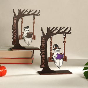 May include: Two wooden Halloween-themed decorations. Each features a tree with a swing, a ghost reading a book, and a library name. One ghost wears a witch hat. The text on the decorations reads "Christy's Library" and "Rowan's Library".