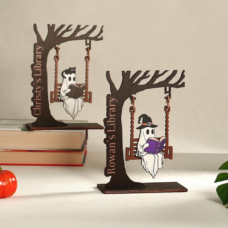 May include: Two wooden Halloween-themed decorations. Each features a tree with a swing, a ghost reading a book, and a library name. One ghost wears a witch hat. The text on the decorations reads "Christy's Library" and "Rowan's Library".