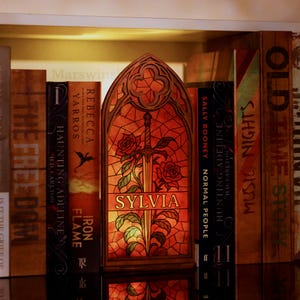Op de afbeelding: Een decoratieve houten boekensteun in glas-in-loodstijl, met een zwaard en rozen, met de naam "SYLVIA". De boekensteun staat tussen andere boeken op een plank, met zichtbare titels.