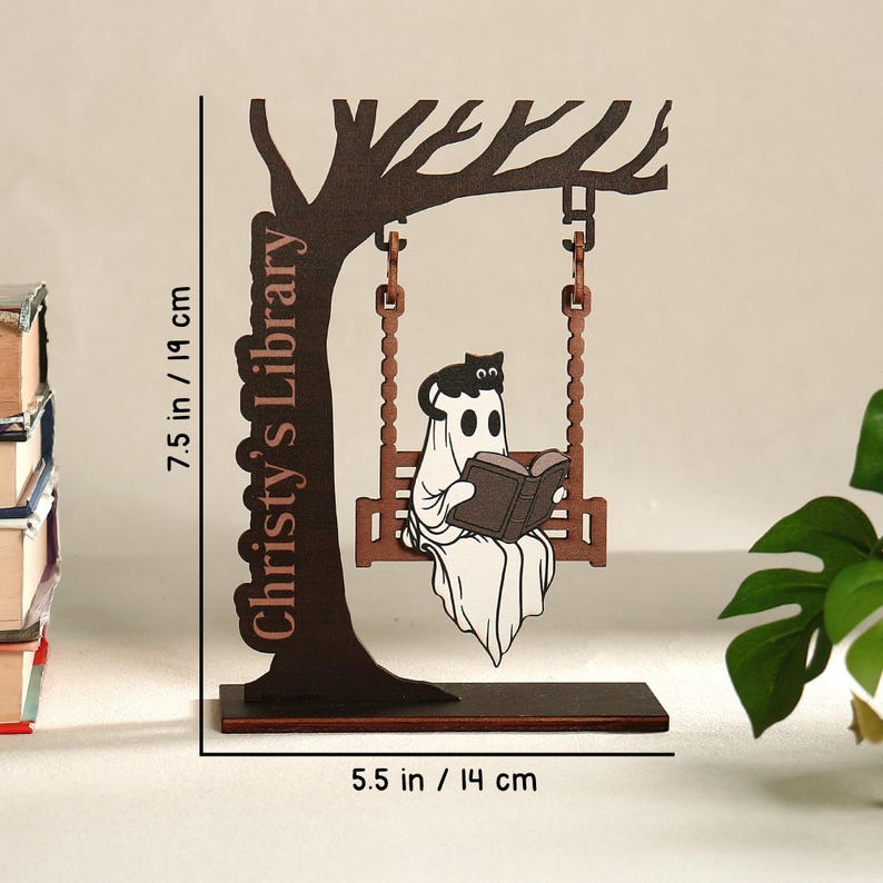 May include: A wooden decorative piece featuring a ghost reading a book on a swing, with a black cat. The tree-shaped design is 7.5 inches tall and 5.5 inches wide. The text "Christy's Library" is on the tree trunk.