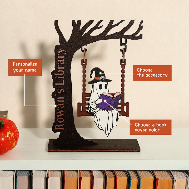 May include: A wooden Halloween-themed bookshelf decoration featuring a ghost reading a book while sitting on a swing. The decoration includes the text "Rowan's Library" and options to personalize the name, accessory, and book cover color.