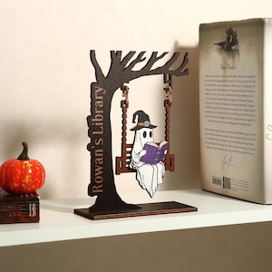 Könnte beinhalten: Eine Holzdekoration zum Thema Halloween, die einen Geist zeigt, der ein Buch liest, während er auf einer Schaukel sitzt. Die Schaukel ist an einem Baum mit dem Text "Rowan's Library" befestigt. Ein kleiner Kürbis und ein Buch sind ebenfalls zu sehen.